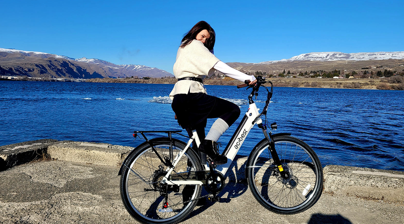 A woman rides a white electric bike on a lakeside path