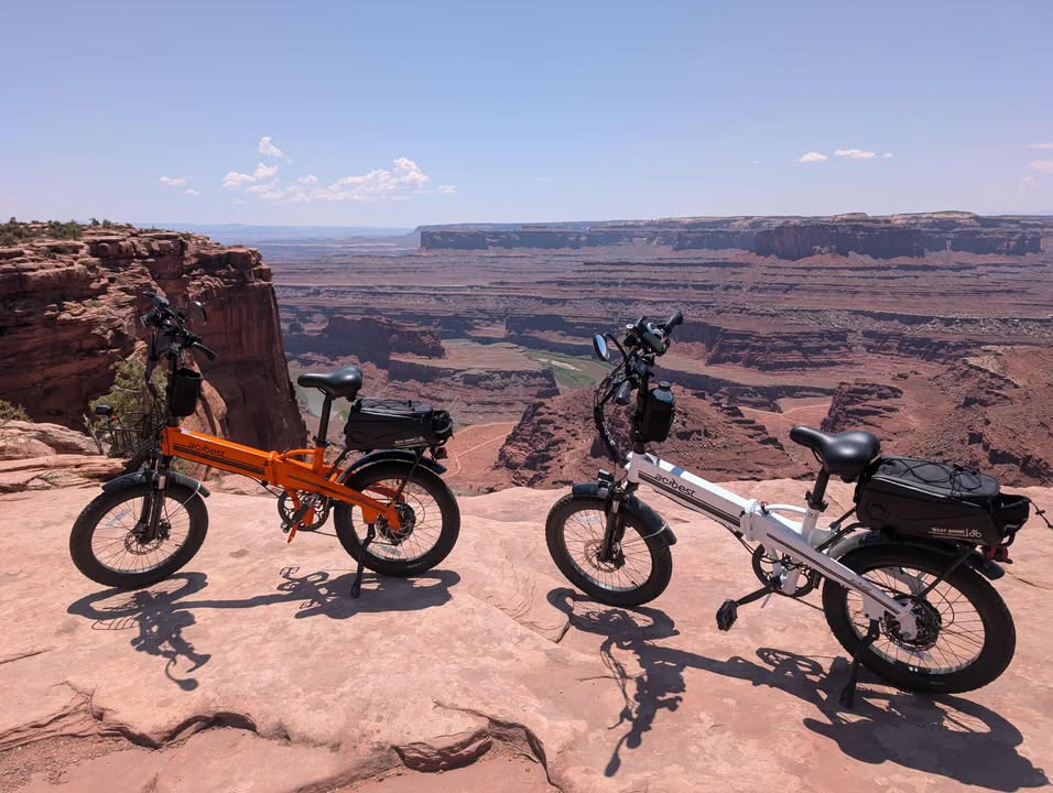 Two electric bikes, one orange and one white, stand on a canyon edge, overlooking vast red rock formations under a clear blue sky.