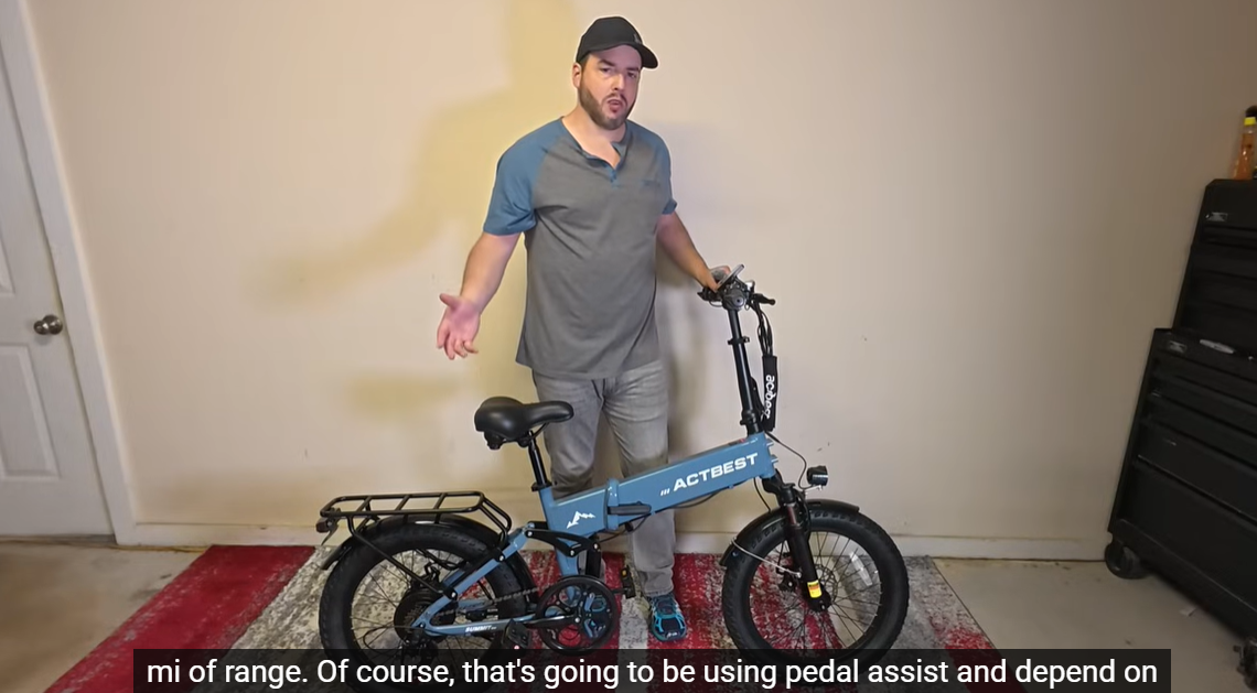 Reviewer standing indoors demonstrating the blue Actbest folding electric bicycle while explaining its range capabilities and pedal assist features