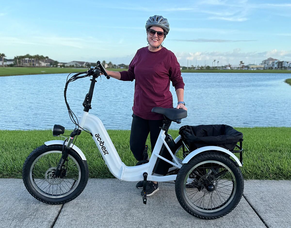 A woman wearing a helmet stands next to a white electric tricycle by a lake.