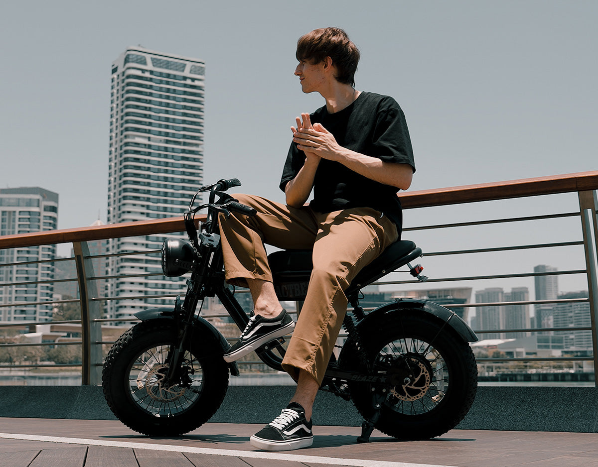 A man wearing a black t-shirt, brown pants, and black-and-white sneakers sits on a black electric motorcycle on an outdoor urban walkway with city buildings in the background.
