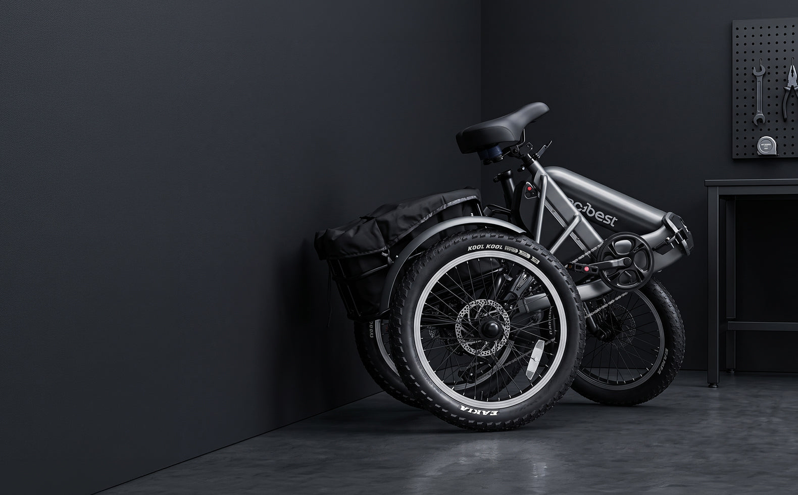 A black and silver electric trike with a rear pannier bag, parked against a dark gray wall in a minimalist studio setting.