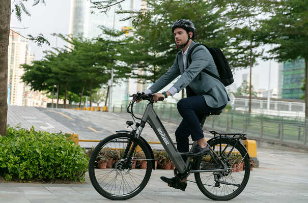 A man in a suit riding a black electric bike, wearing a helmet and carrying a backpack, with a cityscape and green plants in the background.