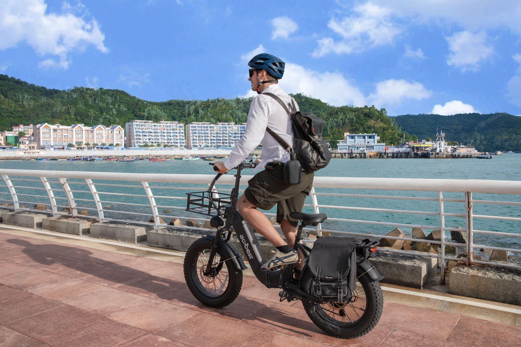Man riding a fat-tire electric bike along a seaside boardwalk, wearing a helmet and backpack, coastal town and hills in the background.