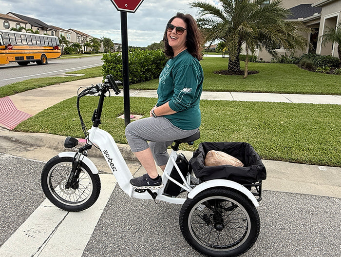 A smiling woman rides a white electric tricycle with a basket on the back.