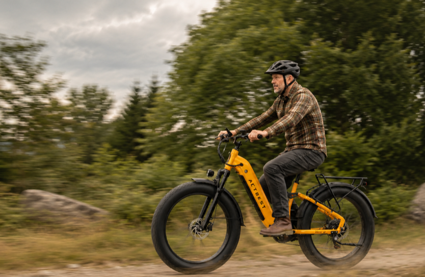 A man riding an electric fat-tire bicycle on a dirt path outdoors, with trees and rocks in the background. The bicycle frame is bright yellow, and the rider wears a helmet, plaid shirt, and dark pants.