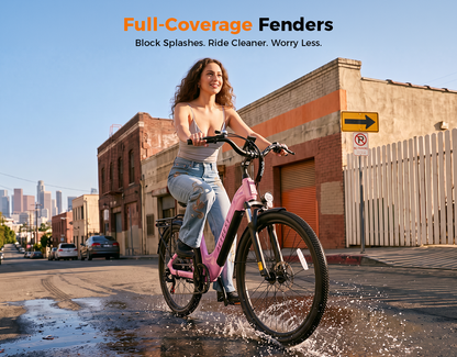 A woman rides a pink electric bicycle through a wet urban street puddle, with buildings, parked cars, and a city skyline in the background. Orange and black text at the top reads 'Full-Coverage Fenders' and 'Block Splashes.