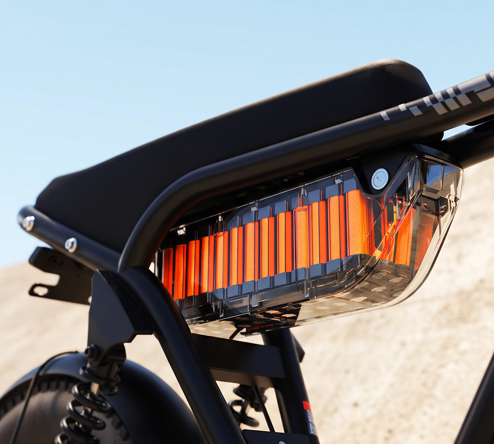 Close-up of a black ebike rear light assembly with bright orange illuminated vertical LED bars under a clear protective cover, mounted on a black frame against an outdoor sandy background.