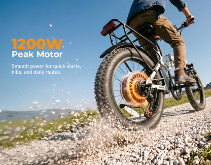 A person riding an electric fat-tire bicycle on a gravel path outdoors under a blue sky. The bike has a large rear tire, a rear rack, and a visible glowing orange rear wheel motor.
