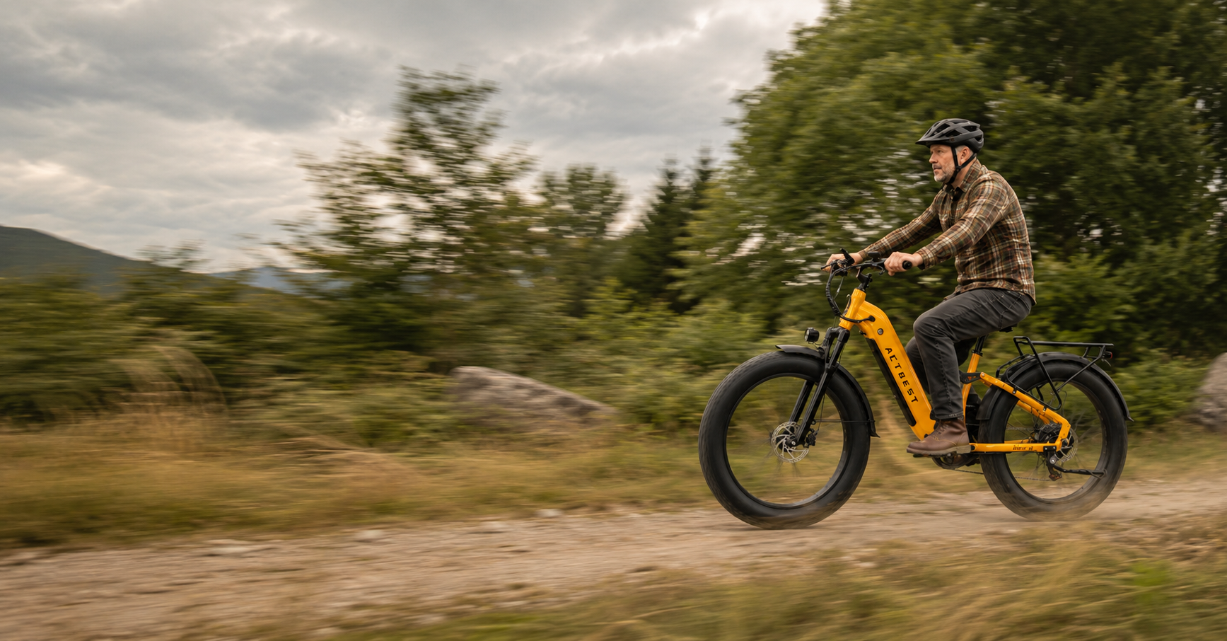 A man riding an electric fat-tire bicycle on a grassy path with trees in the background. The bicycle is bright yellow with black tires and a black rear rack.