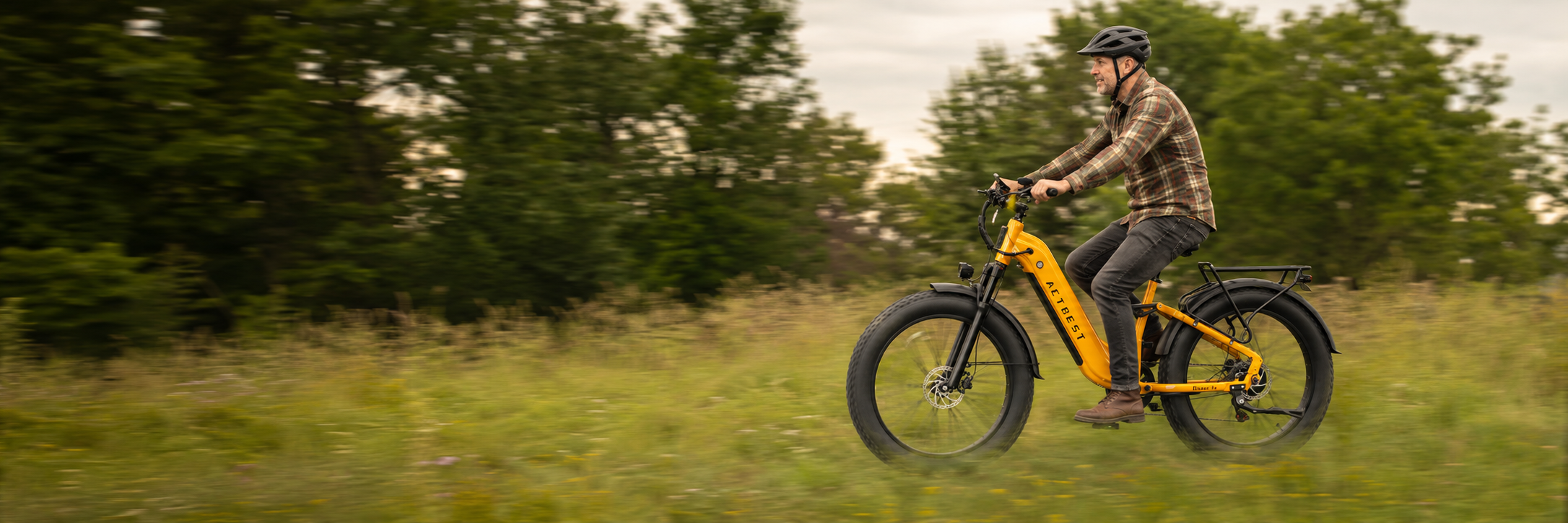A man riding an electric fat-tire bicycle on a grassy path with trees in the background. The bicycle is bright yellow with black tires and a black rear rack.