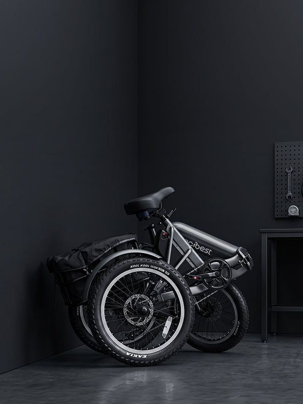 A folded electric trike with a matte black frame and black components, including tires, saddle, and rear rack, parked in a dark studio corner next to a small table.